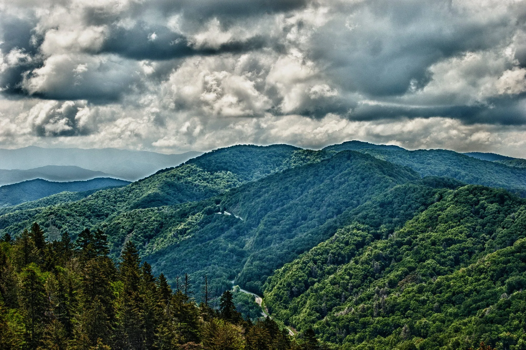 A wide mountain landscape under heavy clouds, with a thin road barely visible winding through the forested valley