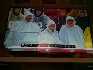 A TV showing three fans in clear rain ponchos in the stands, with the score bar along the bottom