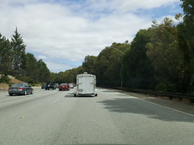 Dashcam view of a multi-lane highway lined with trees, following a small white trailer