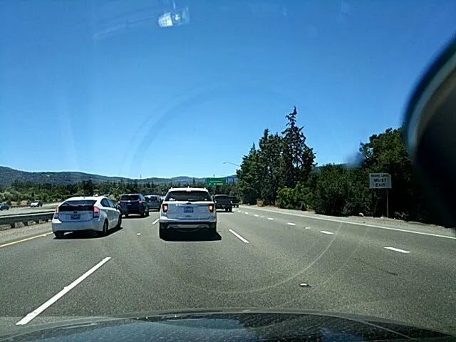 Dashcam view through a windshield of traffic on a sunny highway, with a green exit sign ahead