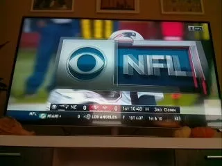 A TV showing a smaller CBS and NFL logo graphic with players visible behind it, with the score bar along the bottom
